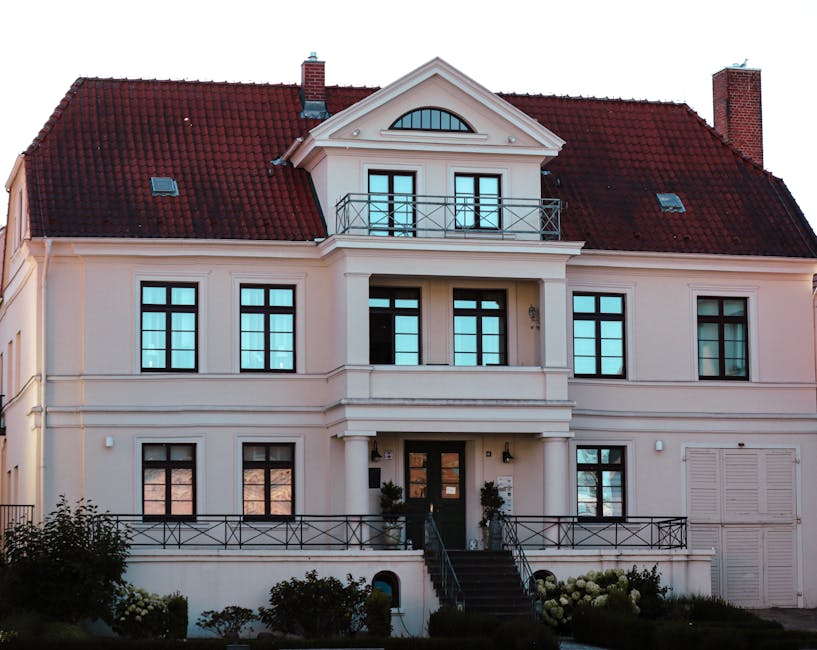 Exterior view of a large, elegant residential building with a cream-colored facade and red-tiled roof, featuring multiple windows with black frames, a central front door with steps leading up to it, and an upper balcony with a metal railing. The well-maintained garden in front has neatly trimmed bushes and flowering plants, illuminated by natural daylight, illustrating the importance of regular exterior cleaning and maintenance services provided by Paddington Cleaners, as referenced in the estate cleaning guide for Sussex Gardens residents.