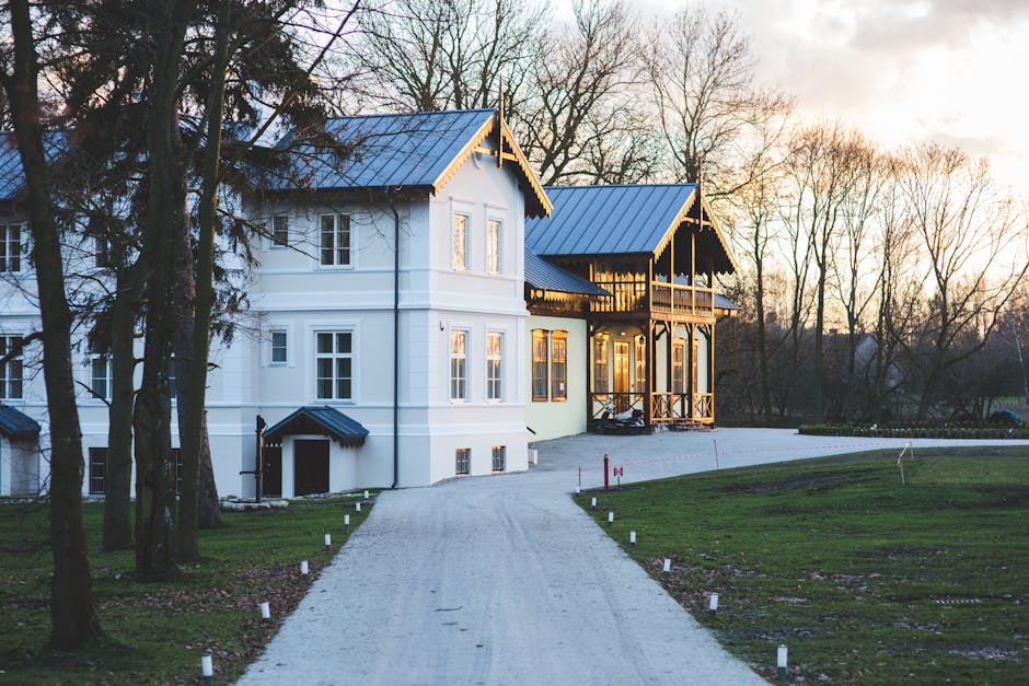 A large, white residential building with a traditional architectural style, featuring a dark-colored roof, multiple double-glazed windows, and a covered porch area. The house is situated at the end of a gravel driveway bordered by small white markers, leading through a well-maintained lawn surrounded by leafless trees. The exterior surfaces of the house appear clean and well-maintained, with visible effort in cleaning and upkeep, indicative of professional cleaning services. The warm lighting from inside the house emits a welcoming glow through the windows, highlighting the building's neat and tidy appearance, consistent with standards of domestic cleaning and surface sanitisation. The overall scene is captured during a sunset or early evening, providing soft natural lighting that enhances the exterior's pristine condition, aligning with the principles of deep cleaning and maintenance promoted by Paddington Cleaners for Sussex Gardens residents.