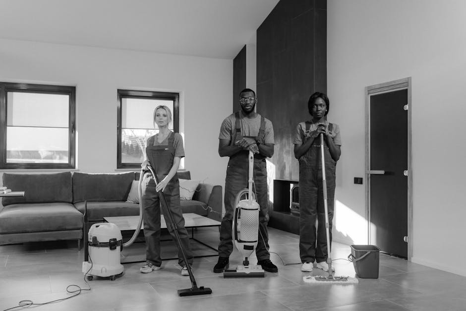 Close-up view of three professional cleaners dressed in gray shirts and bright orange overalls, standing indoors on a light-colored tiled floor. The cleaners are holding cleaning tools; the central wo